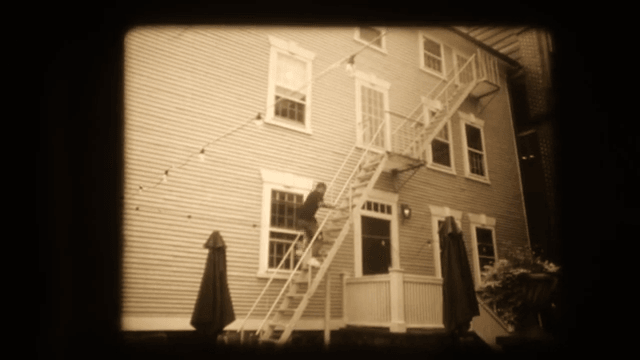 16mm frame of a house with an external white staircase and two figures in dark coats